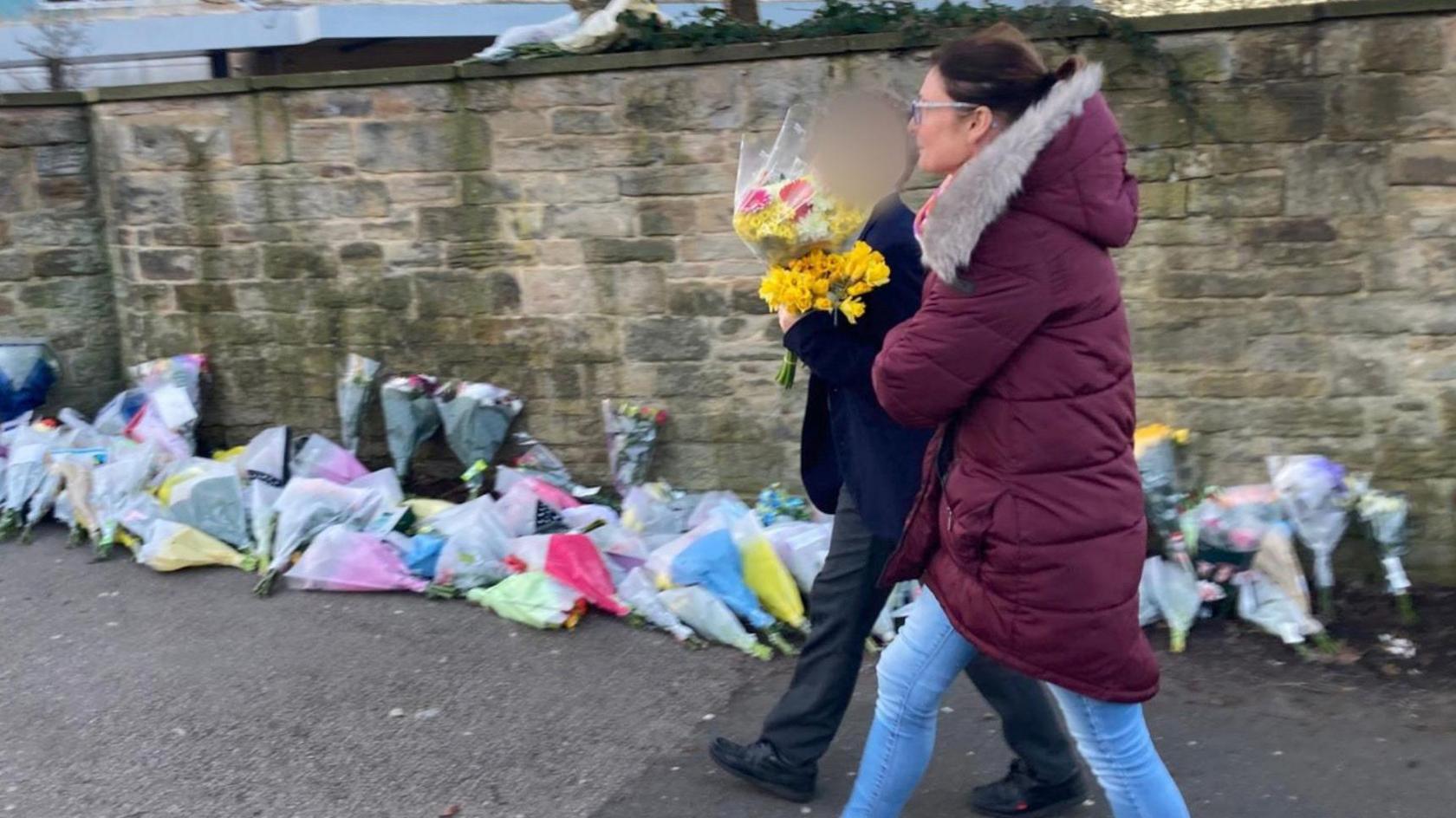 A pupil holding a bouquet of flowers as they walk past a wall lined with floral tributes.