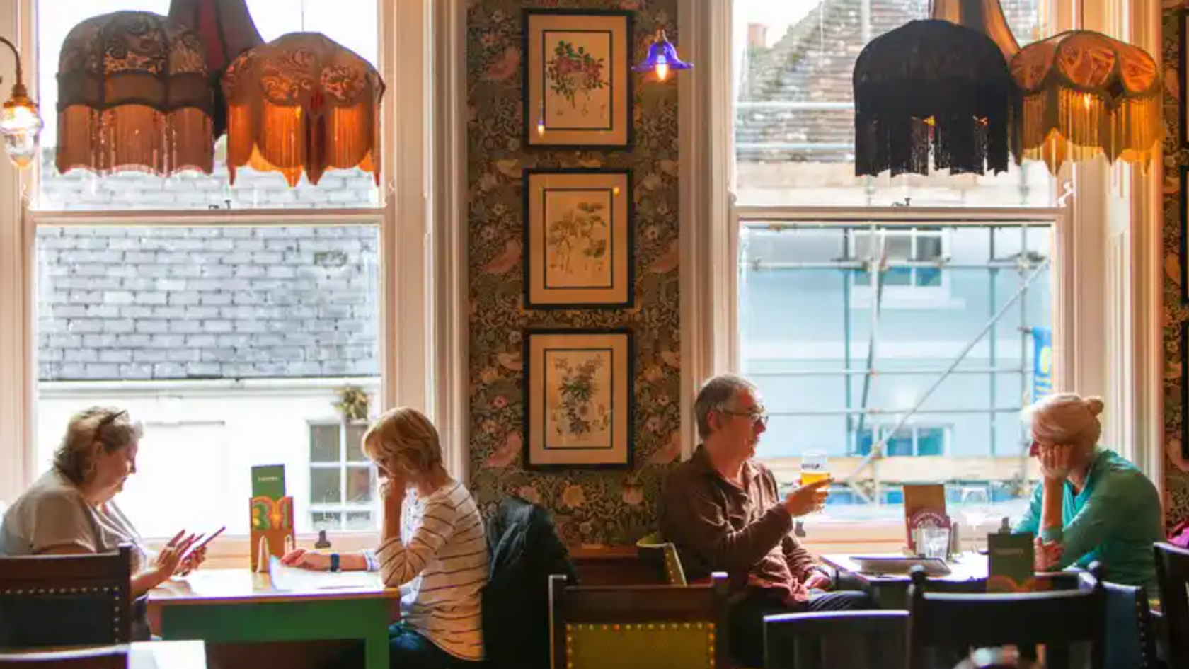 The interior of a Lounge restaurant in Warminster. There are two couples sitting at separate tables, surrounded by floral wallpaper and framed prints. There are various colourful lampshades with tassels hanging above. 