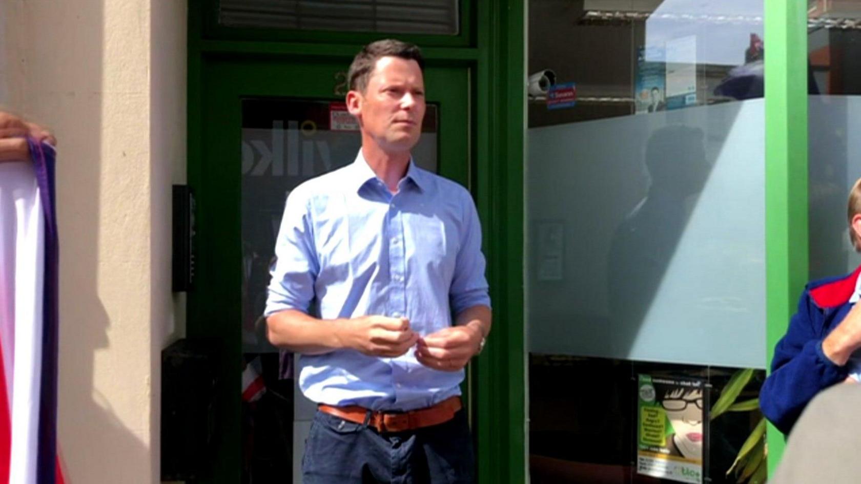 Alex Chalk standing in a doorway wearing a pale blue shirt and dark trousers adressing a crowd of protestors with a grave look on his face