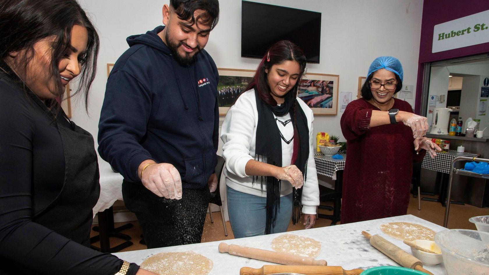 A young man and three women sprinkle flours over dough on a white table with rolling pins