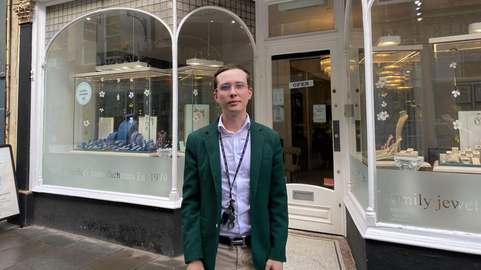 Charlie Dalton-Ball has dark short hair and is wearing glasses, a dark green blazer over a shirt and beige chinos. He is standing outside of his business which has large display windows and white painted wooden panelling.