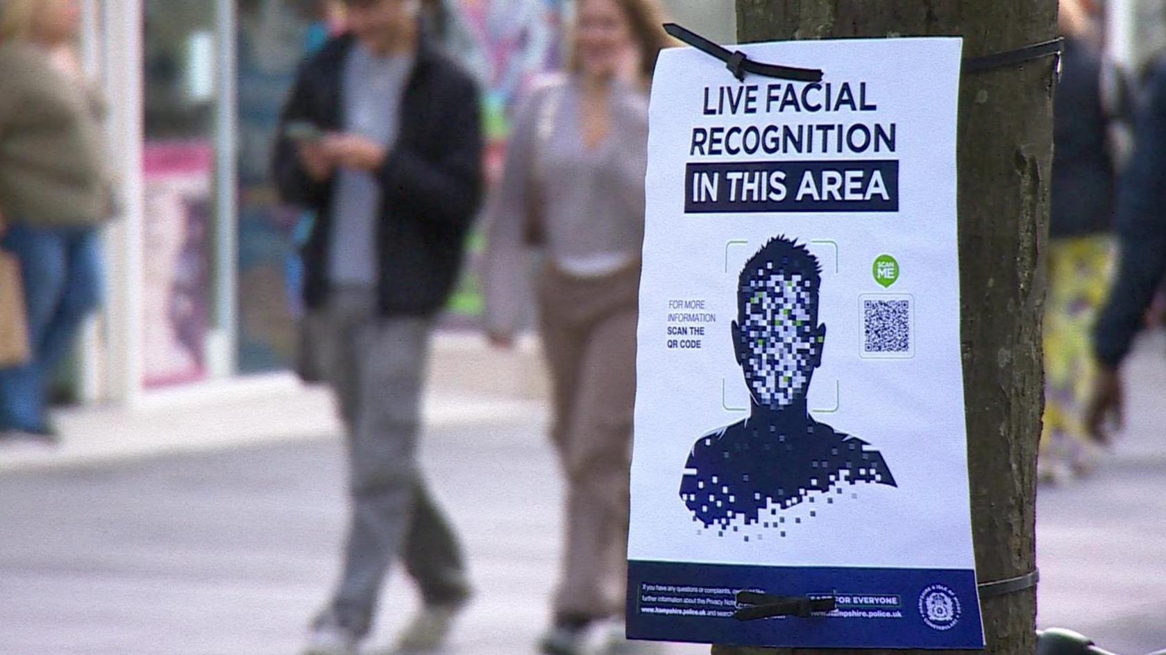 A sign posted to a tree saying 'facial recognition in this area' as people walk past in a street behind it