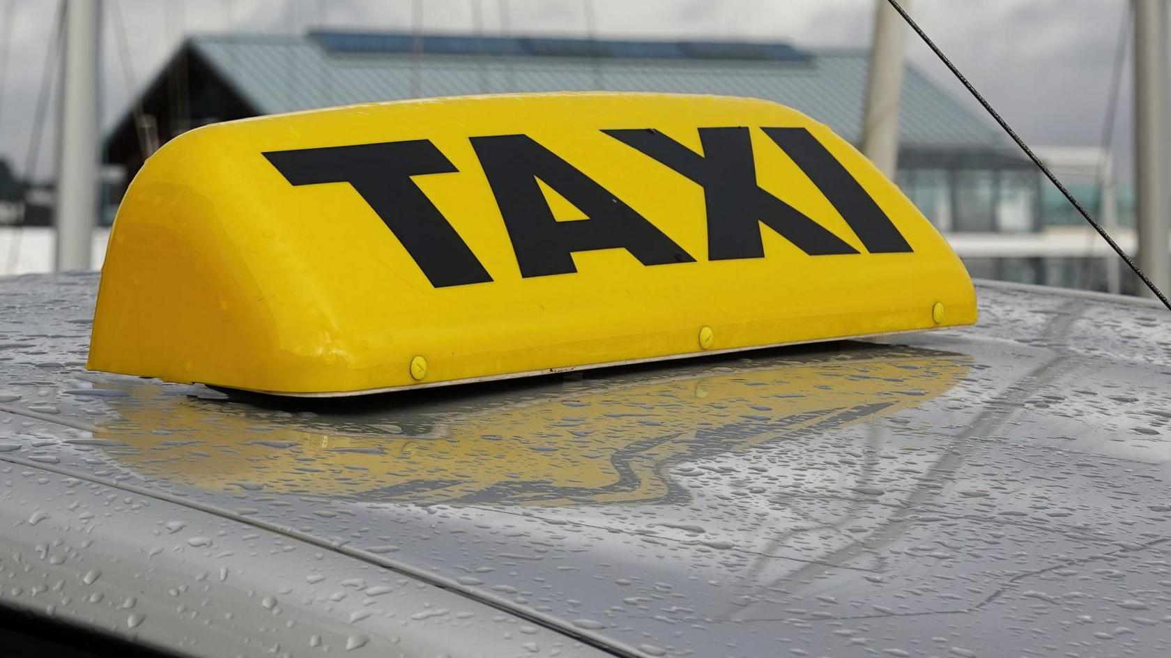 An unlit yellow taxi light on the top of a car on a grey, wet day in the British Isles.