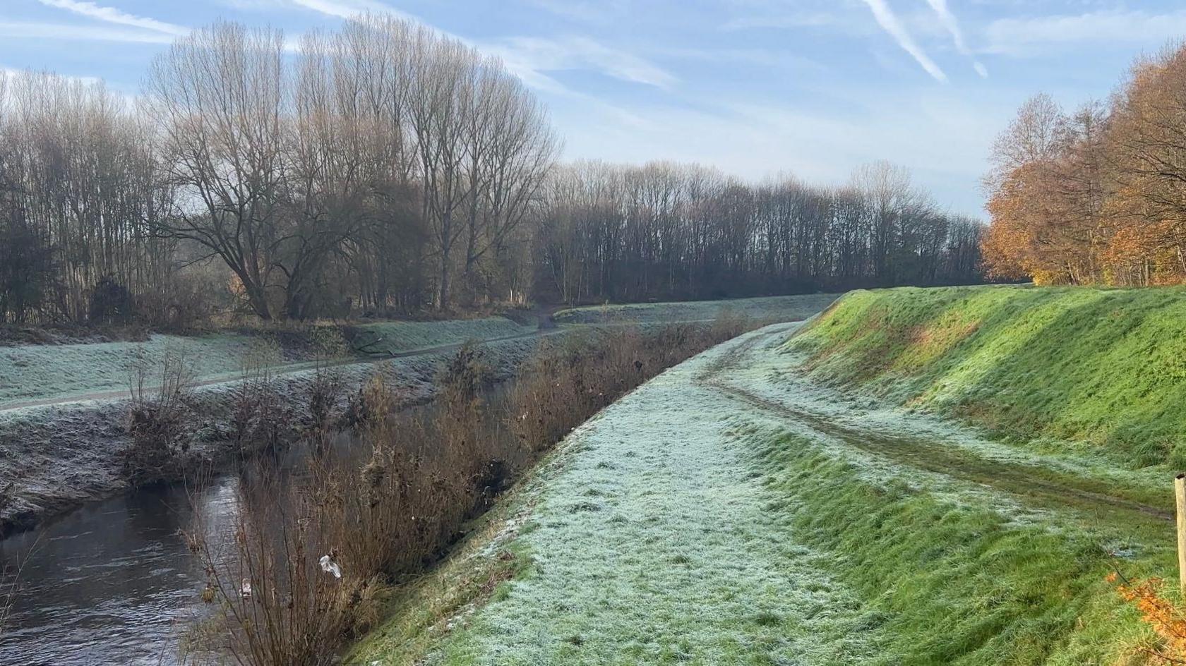 The banks of the River Mersey, surrounded on one side by a sloped, grass walkway, parts of which have frosted over, and on the other, another grass verge with trees lining the scene. 