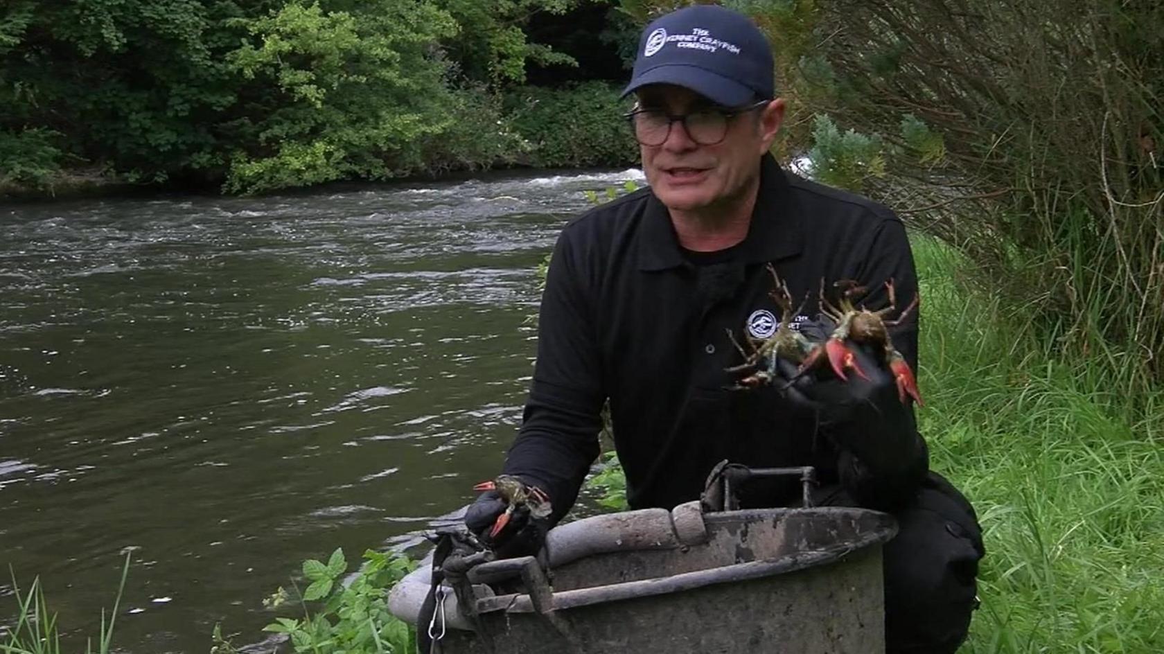 Andrew Leech crouches on a riverbank behind a large bucket, he is holding a live crayfish, wearing a black polo shirt with a black long sleeved shirt underneath and black trousers, he has a dark blue peaked cap on with the logo for the Kennet Crayfish Company