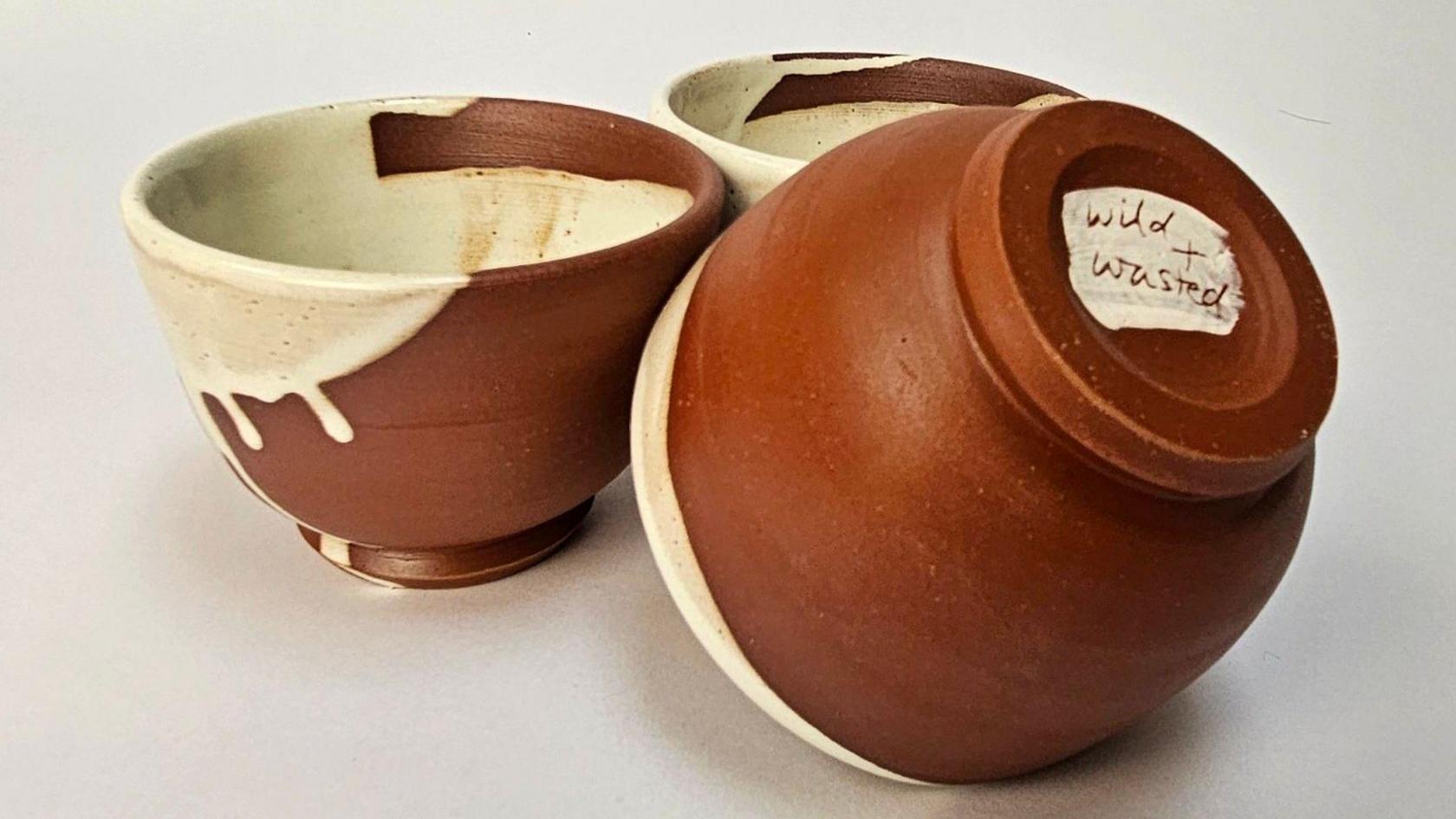 Three terracotta coloured bowls with a dripping cream glaze covering part of the bowls - one is overturned and says Wild and Wasted on the bottom of it