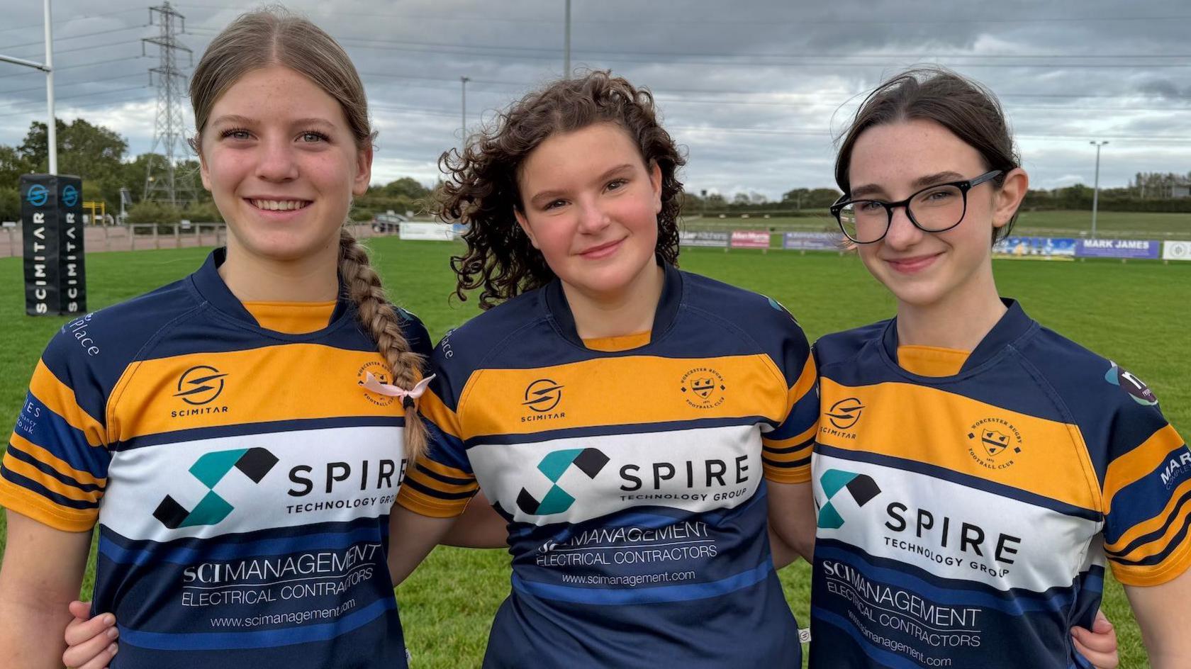The three teenagers are standing on a rugby pitch with their arms around each other.  They are smiling and wearing blue and gold Worcester rugby club shirts. 