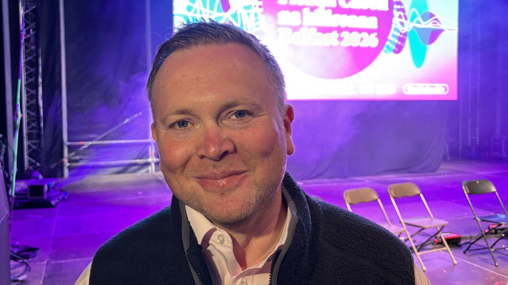 Maurice has short spiked hair and wears a fleece bodywarmer and shirt. Behind him is the main stage of the fleadh, where a few fold up chairs are bathed in purple light. 
