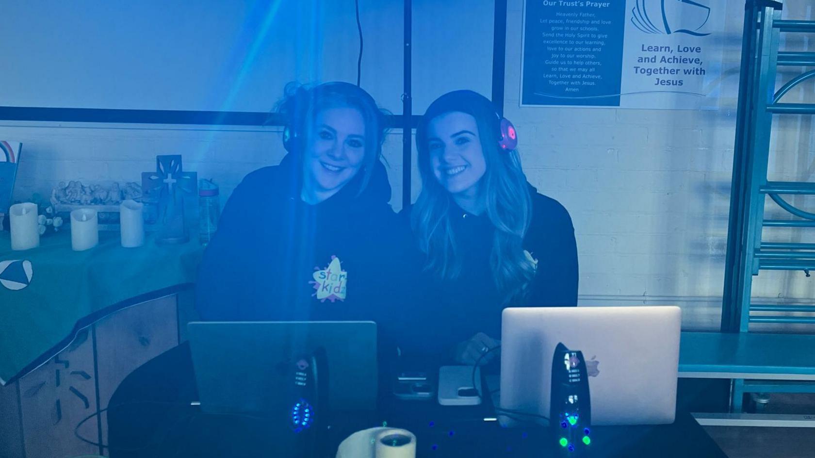 Jan and Ellie Hayes smile while sitting behind the mixing desk at the silent disco. They have computers in front of them and are wearing headphones.