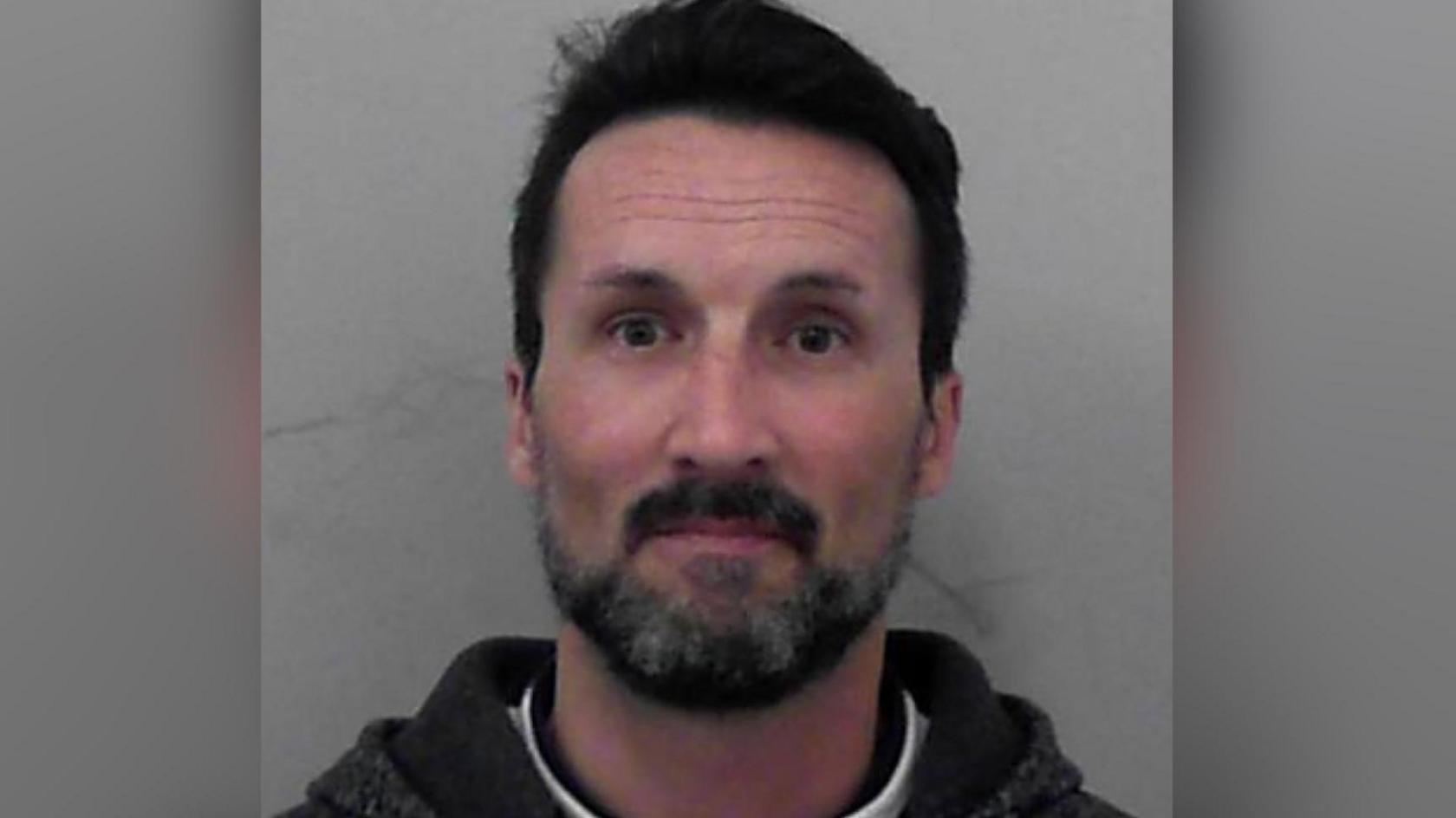 A mugshot of Mark Acklom. He has short dark hair and facial hair. He is pictured against a plain background with a blank expression. He is looking directly at the camera.