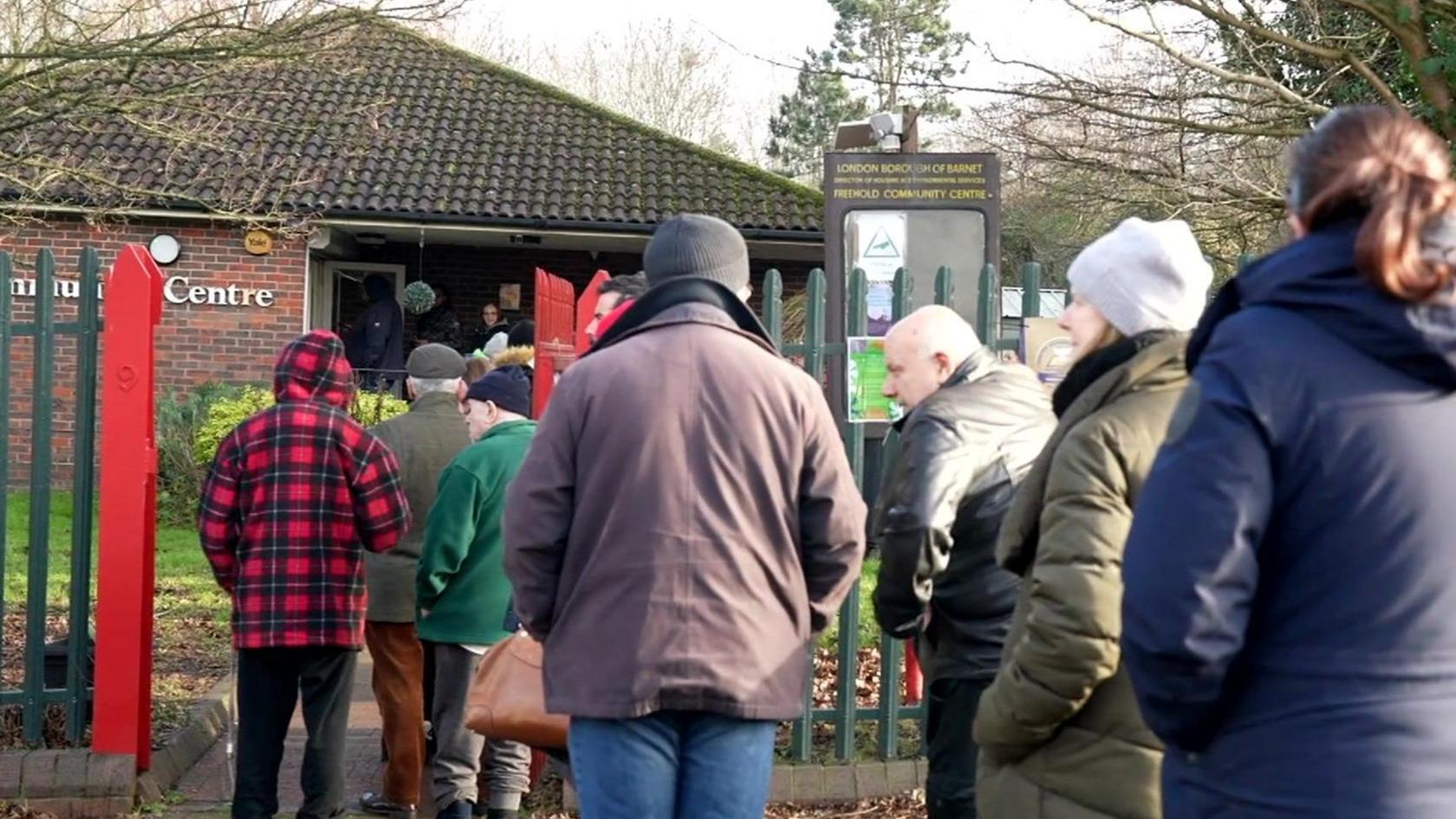 A group of people wearing coats queuing outside a brick building.