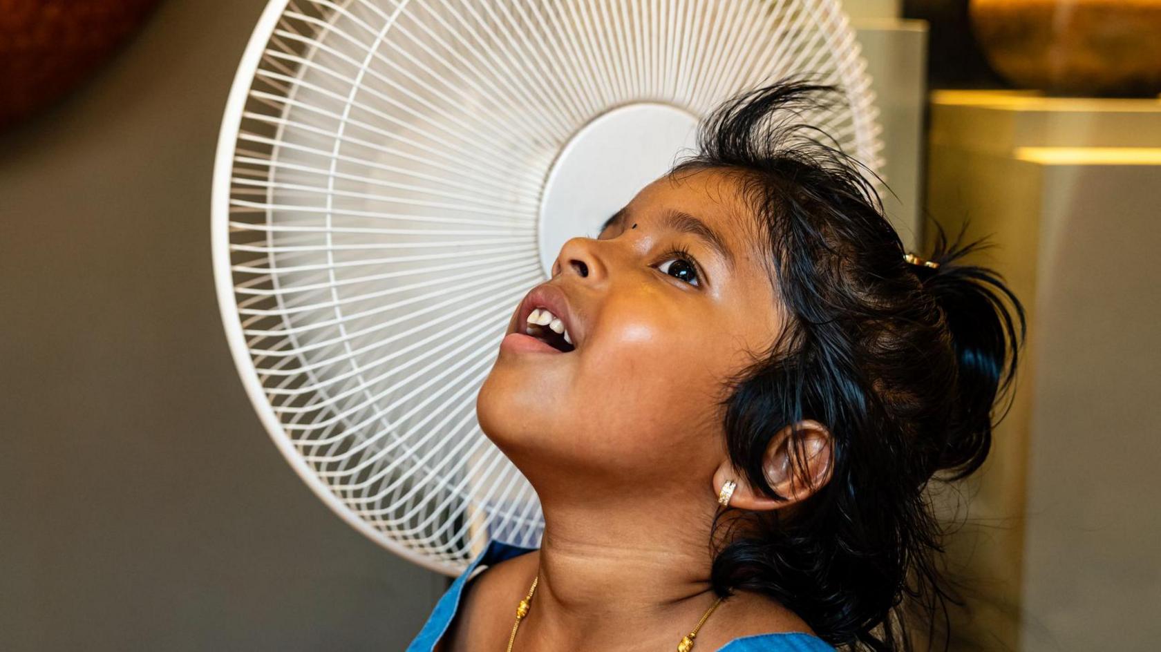 A little girl stands next to a whirring fan.