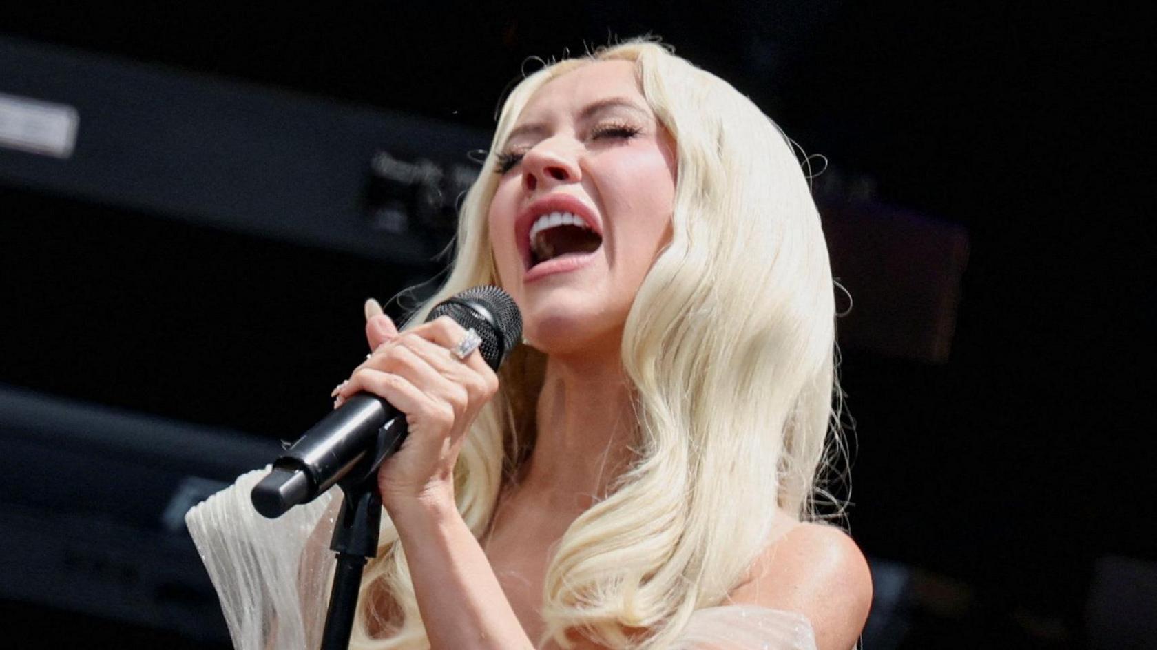 Christina Aguilera is pictured with her eyes closed as she sings on stage. She has light blonde, long, wavy hair and wears a white dress.