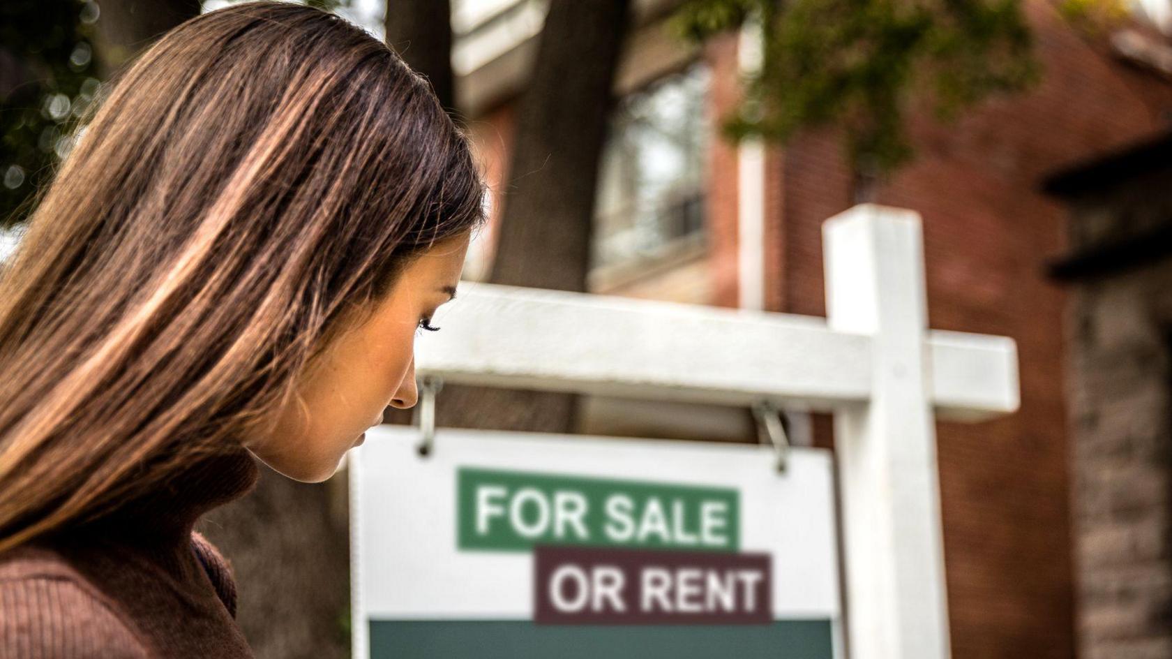 Woman looking toward house with for sale sign in front of it