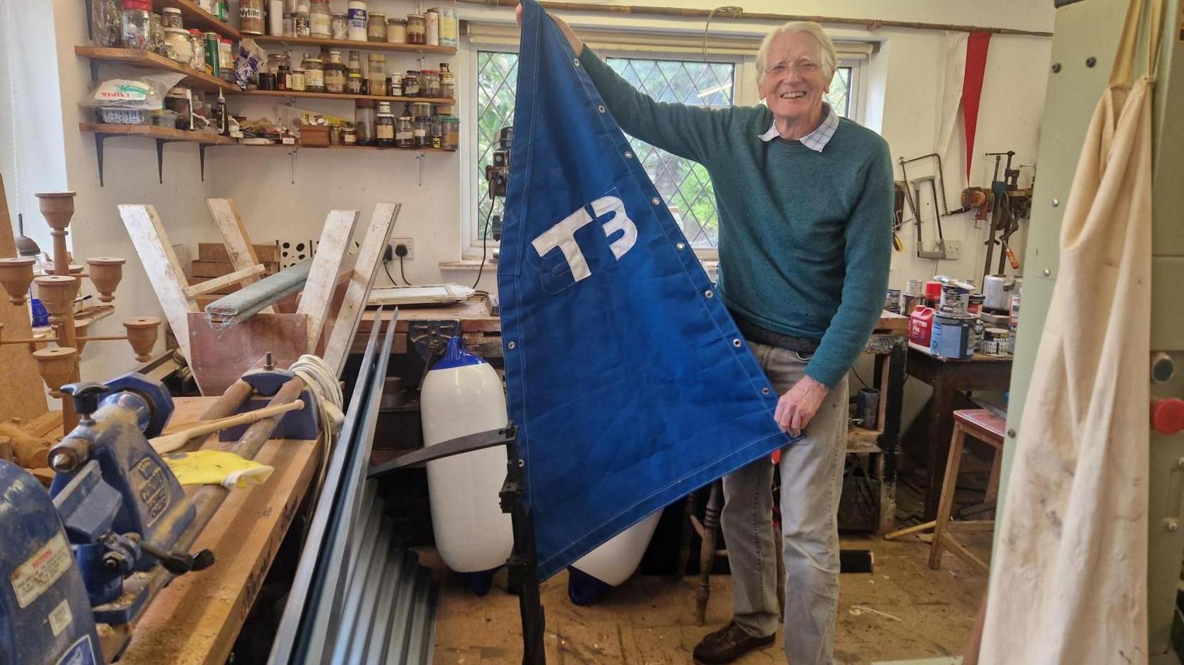 An elderly man with white hair holding a blue sail with T3 on it in white. He is standing inside a workshop with tools and differet materials around including corrugated iron, wood and metal