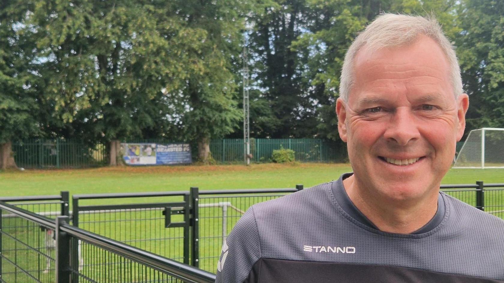 A man in a grey football shirt standing by the edge of a football pitch, leaning against a metal fence. There's a banner saying Bearsted FC in the background.