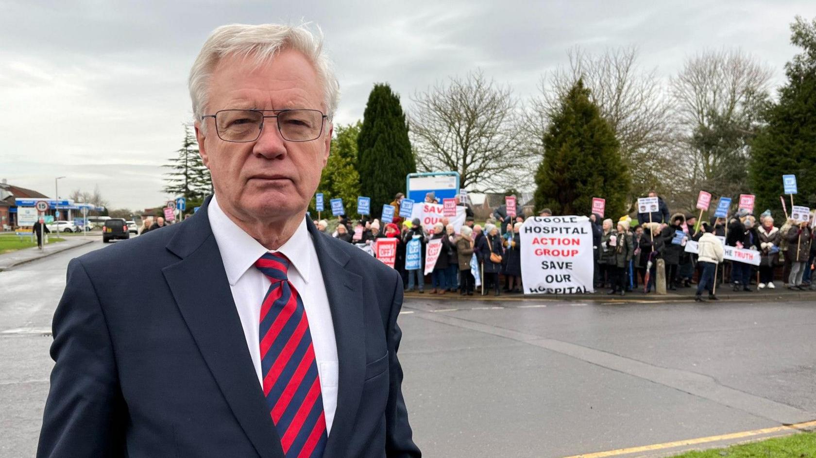 MP Sir David Davis talking to BBC Look North. He is wearing a dark-coloured suit and a red striped tie. Campaigners can be seen holding placards in the background.