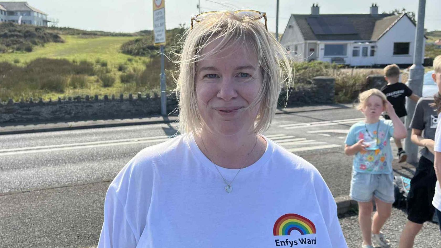 Catrin Elin smiles at the camera. She has short blonde hair, with a pair of glasses resting on the top of her head. She is wearing a white t-shirt with a rainbow logo and the words Enfys Ward beneath. Behind her is a road, with some children visible to her right.