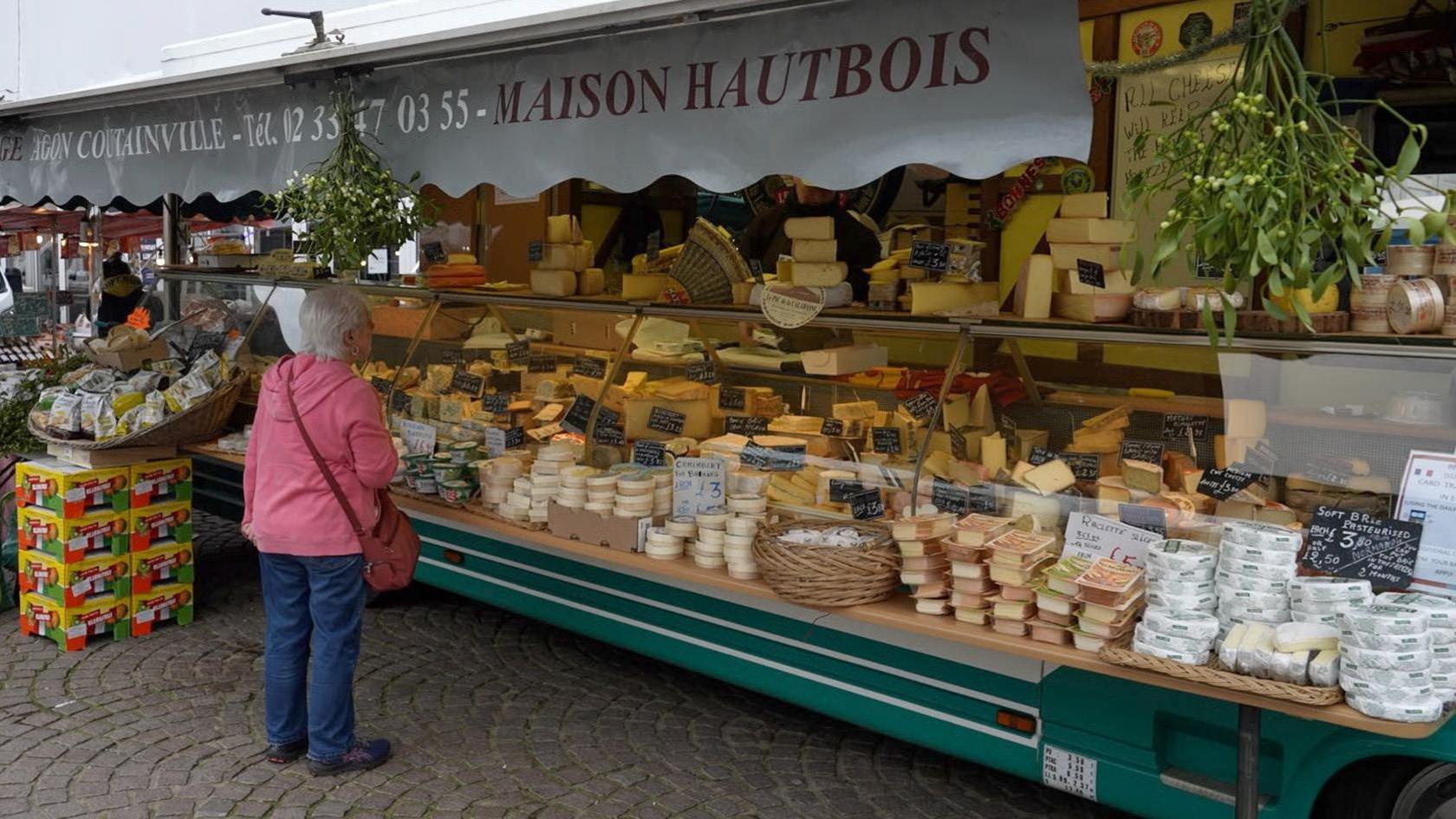 A woman stands in front of a large glass-fronted counter that is full of many different types of cheese. The banner above the counter reads MAISON HAUTBOIS and there is also a bunch of mistletoe hanging.