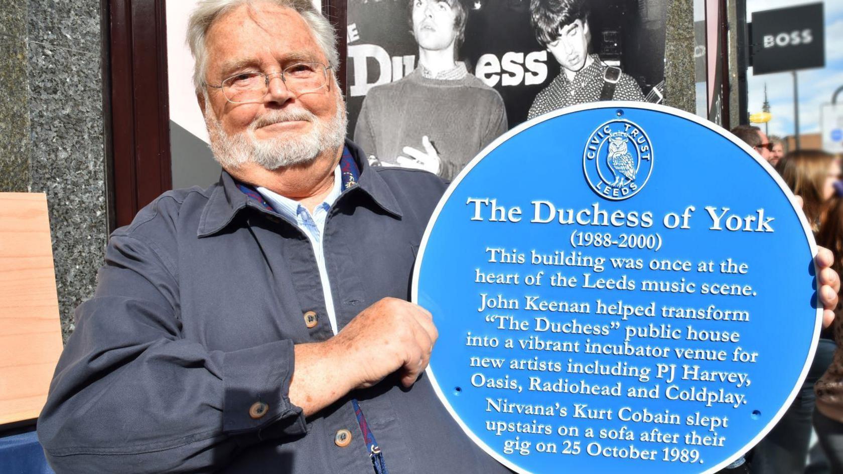 A man with grey hair and a beard is wearing glasses and a navy jacket and holding up a blue plaque, which says The Duchess of York. He is standing in front of a black and white image of Oasis members Liam and Noel Gallagher when they were younger, on stage at The Duchess of York.