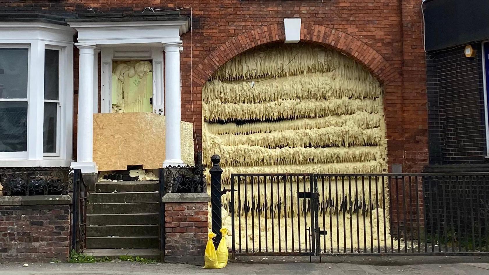 A close up of the entryway and archway of a brown brick house. Both are filled with light yellow foam that has solidified and dropped. The doorway has two pillars outside it, and there is a black metal gate in front of the entryway to the arch