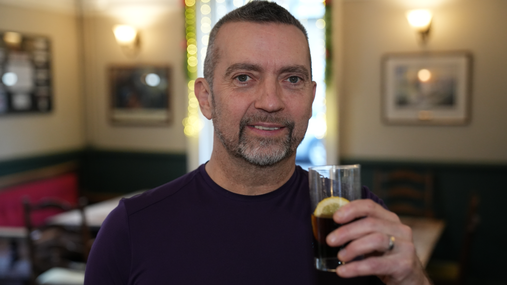 A man in a purple top holding a drink in a glass with a wedge of lemon. He is standing in a pub.