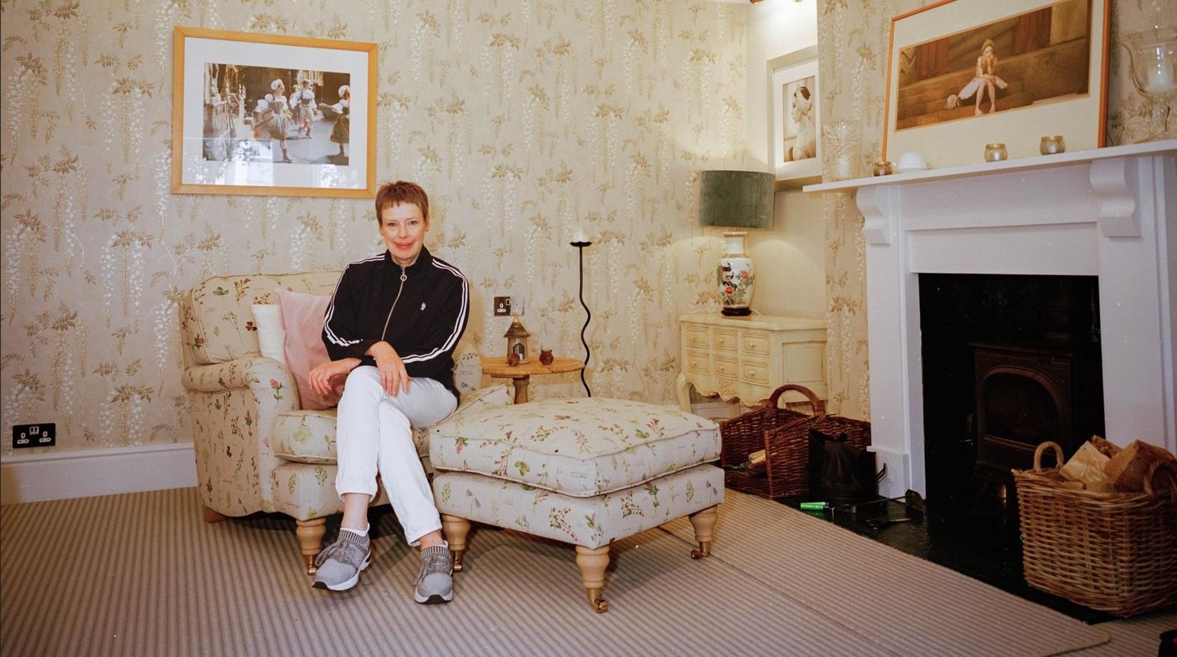 A woman wearing a tracksuit jacket sat on a floral printed arm chair and footrest. She is in a living room that has a few paintings on the wall, floral wallpaper and a black fireplace to the right.