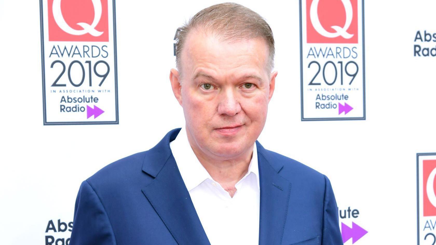 Edwyn Collins standing on the red carpet at an awards ceremony. He is wearing a blue suit with white shirt and has brownish hair.