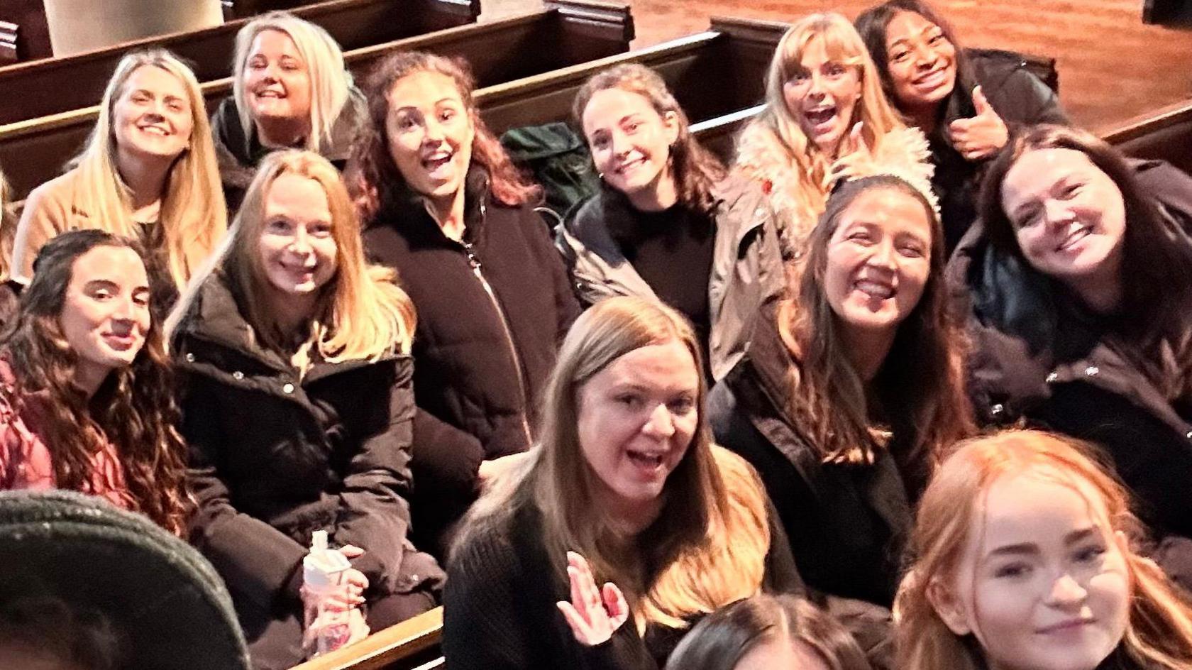 A group of women sit in church pews, they are all smiling at the camera with some doing a thumbs up sign. 