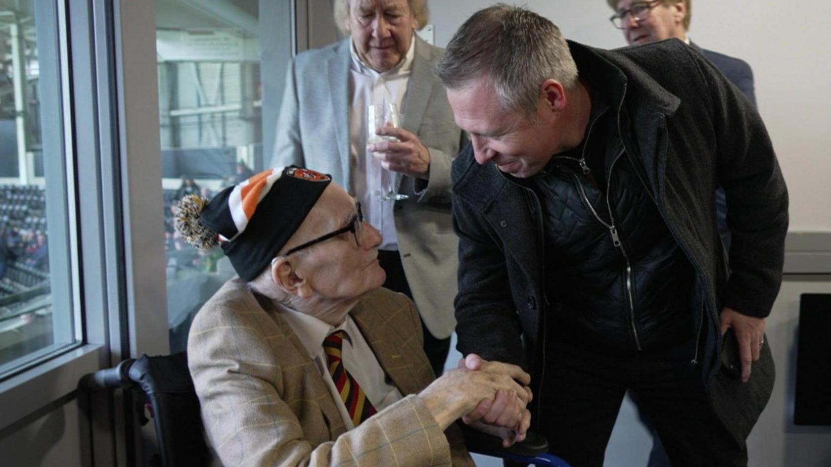 Noel Hebbs, a 100-year-old man shakes the hand of former footballer Nick Barmby in an executive box at a football stadium as his two sons look on. Mr Hebbs, who wears glasses, is sitting by a window and dressed in a smart jacket, Hull City tie and Hull City hat. Mr Barmby, who has short grey hair, is dressed in black and smiling as he looks down at Mr Hebbs.