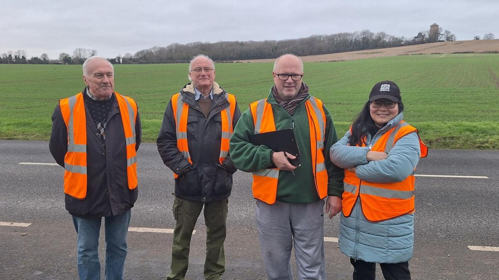 Three men and one woman are stood at the side of a road in front of a large green field. They wear orange high visibility vests and are looking at the camera. One member is carrying a clipboard and pen.