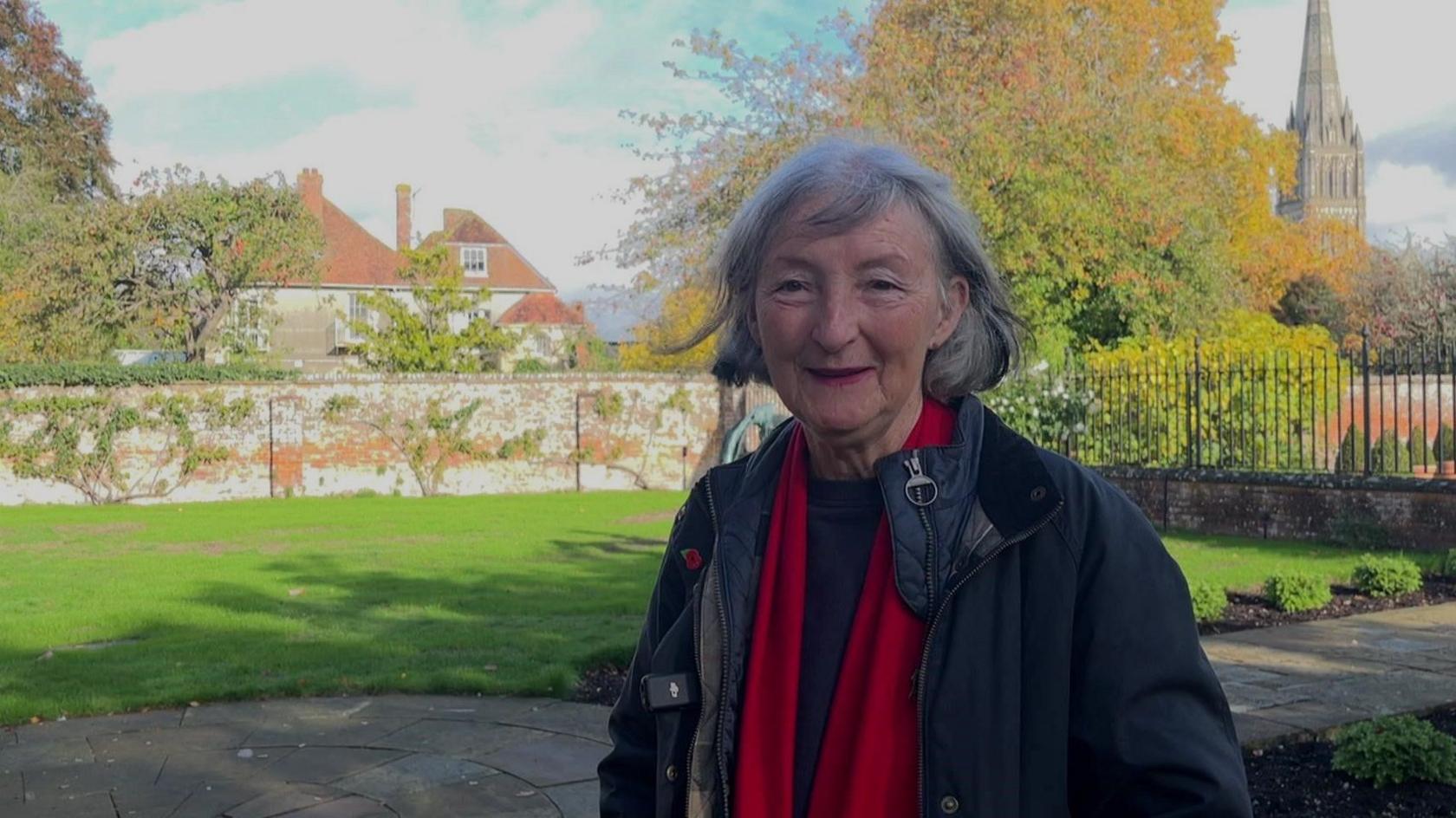 Liz Salter, from Salisbury Cathedral Close Preservation Society, stands in the garden of a neighbouring property to Leaden Hall. Behind her right shoulder can be seen the terracotta coloured roof of the Georgian house and over her left shoulder the spire of Salisbury Cathedral. The brick wall surrounding Leaden Hall can be seen adjoining the lawn of the garden.