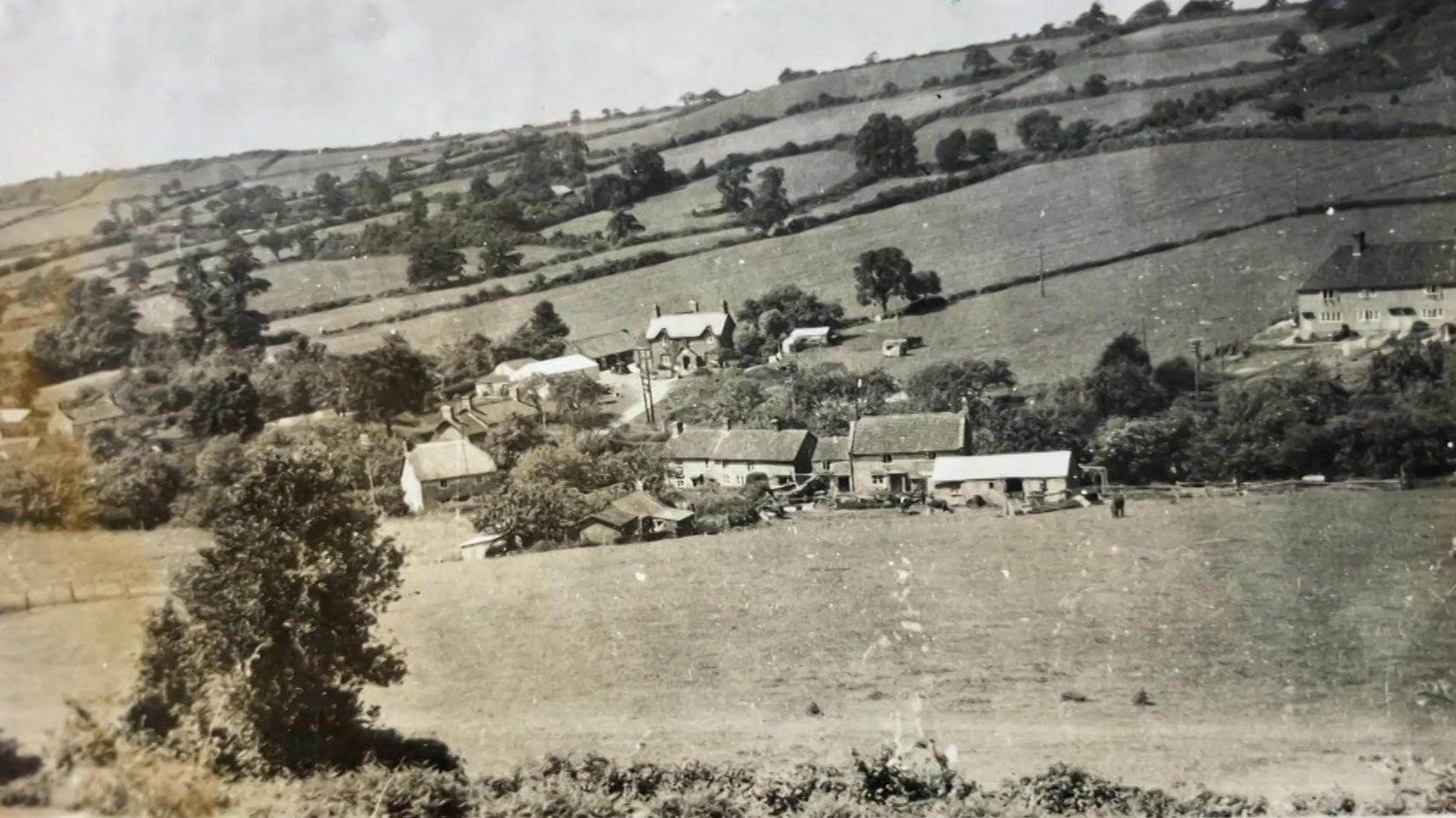 An historic photograph of Hardington Mandeville village, Somerset. There are acres of fields and hedgerows with a round five or six buildings and outbuildings in the middle of the picture, which could be a mixture of homes and the school. 