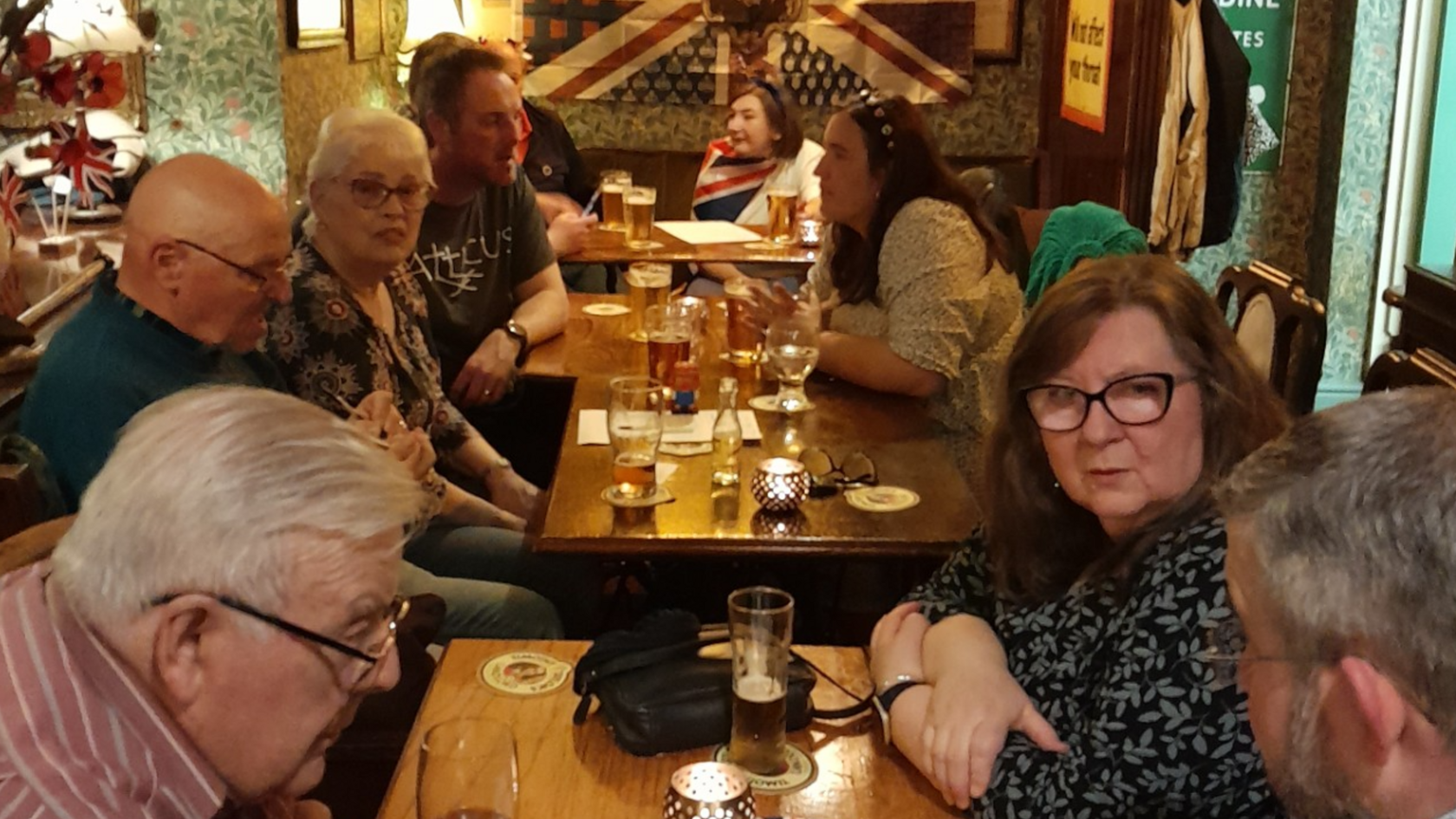 People huddled around tables at the Albion Inn drinking together and talking. In the background on the wall there is a Union flag and old memorabilia from the early 20th century.