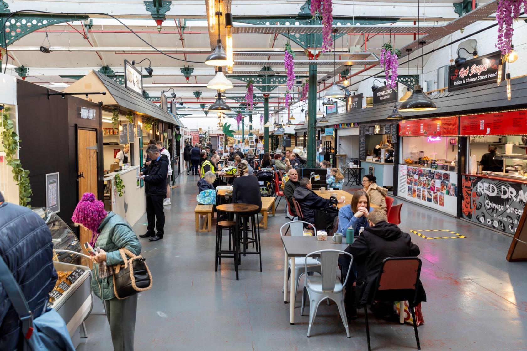 An indoor market lined with stalls on either side and tables and chairs in the centre. The markets is busy with people browsing stalls and eating.