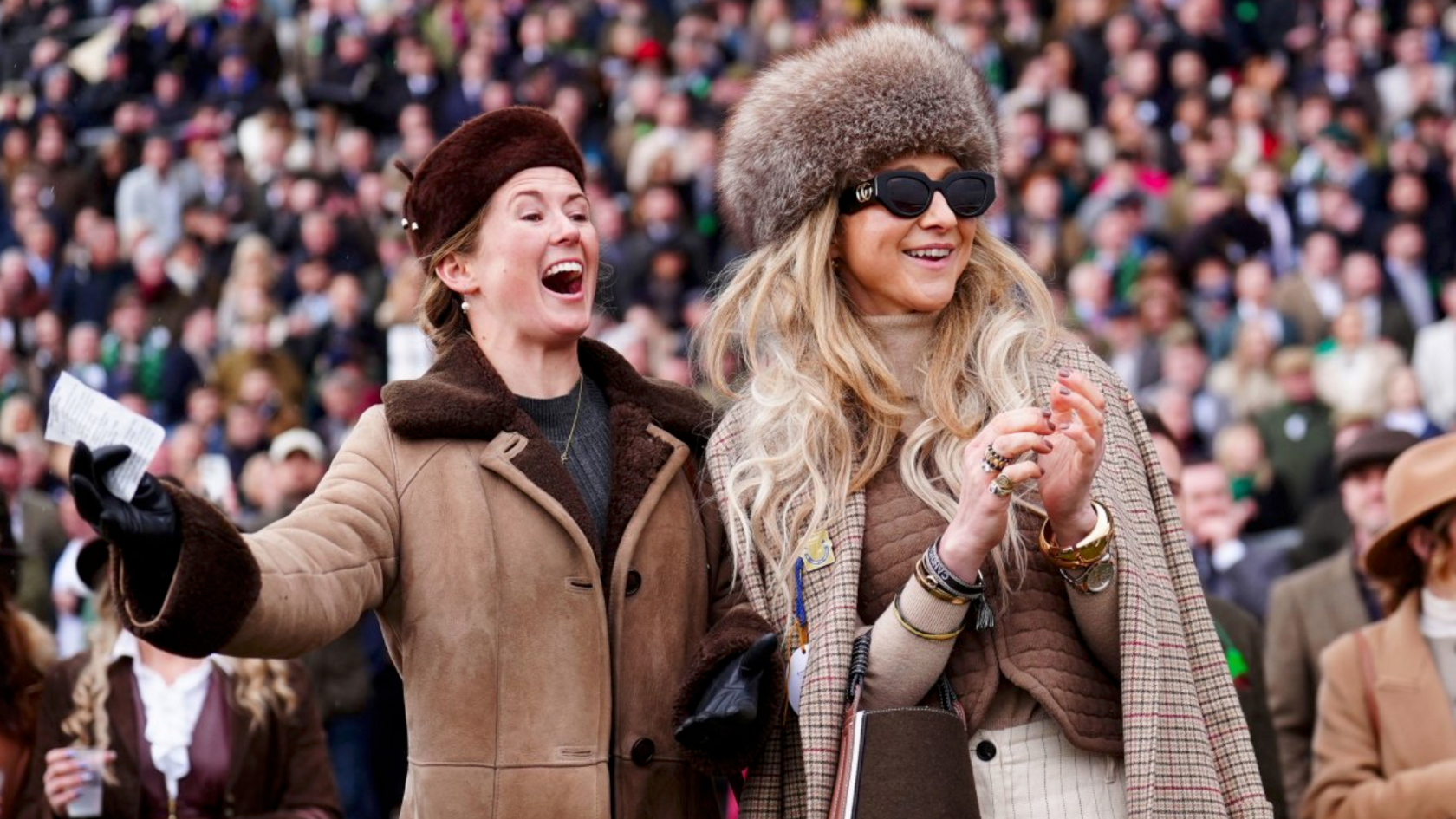 Two female spectators dressed in brown and cream outfits look to the right and cheer as they watch a race.