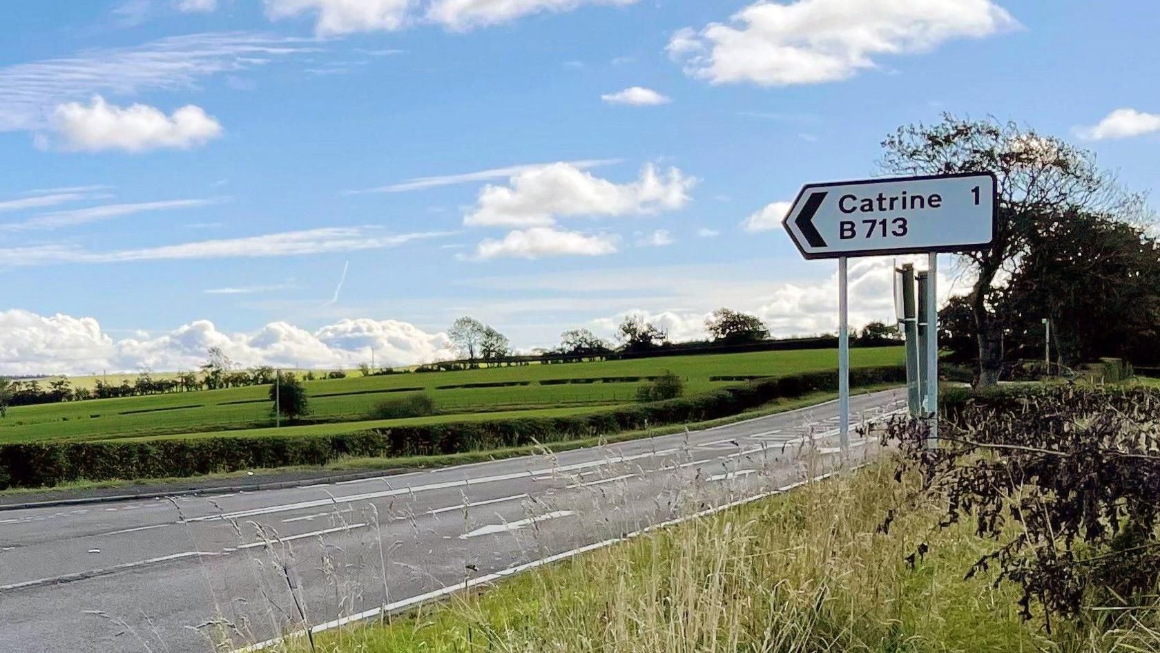 Road sign in grass at side of road with blue sky and fields in background