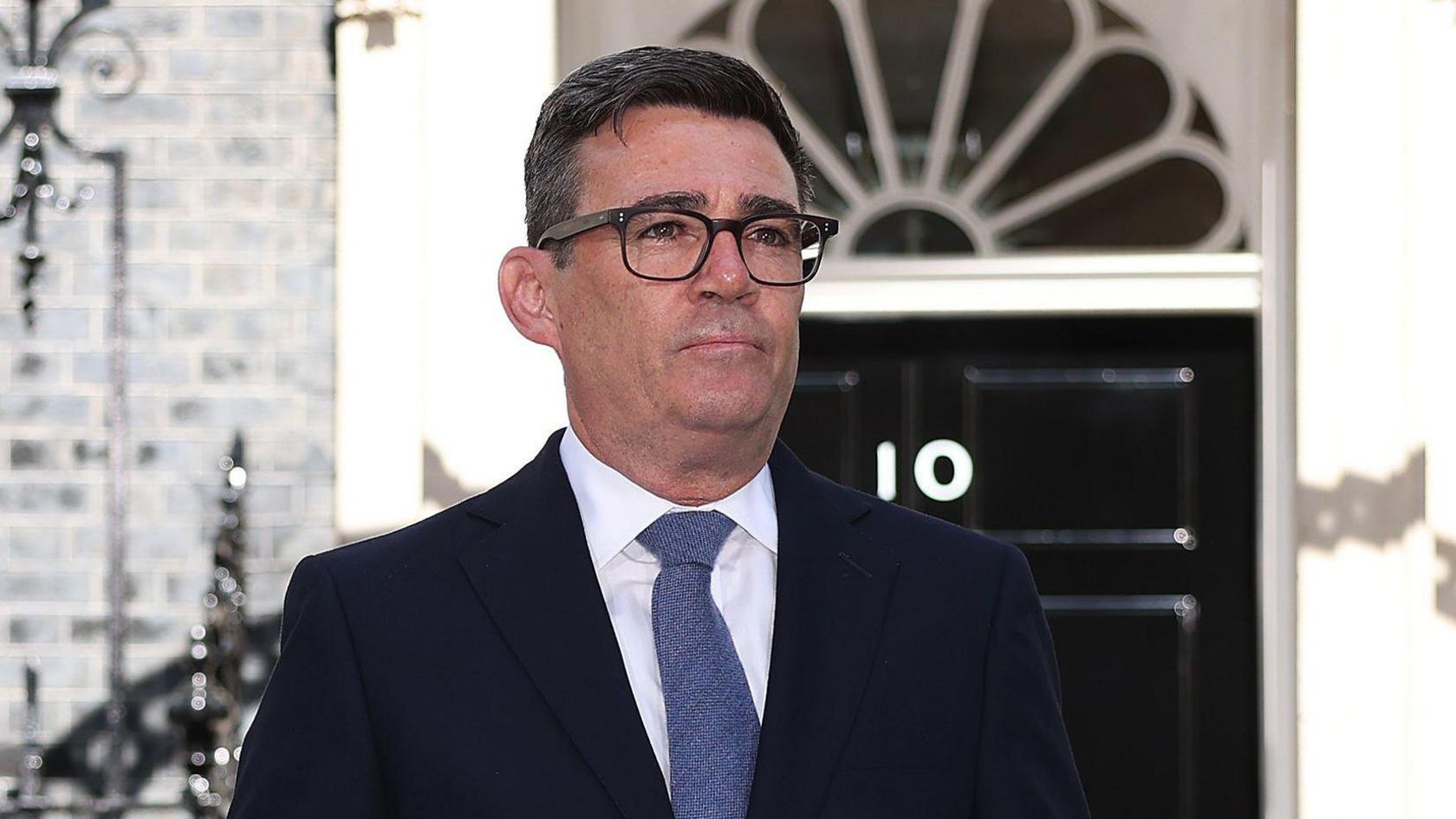 Andy Burnham has short dark hair, wears dark glasses and a dark suit with grey tie. He stands outside the front door of 10 Downing Street