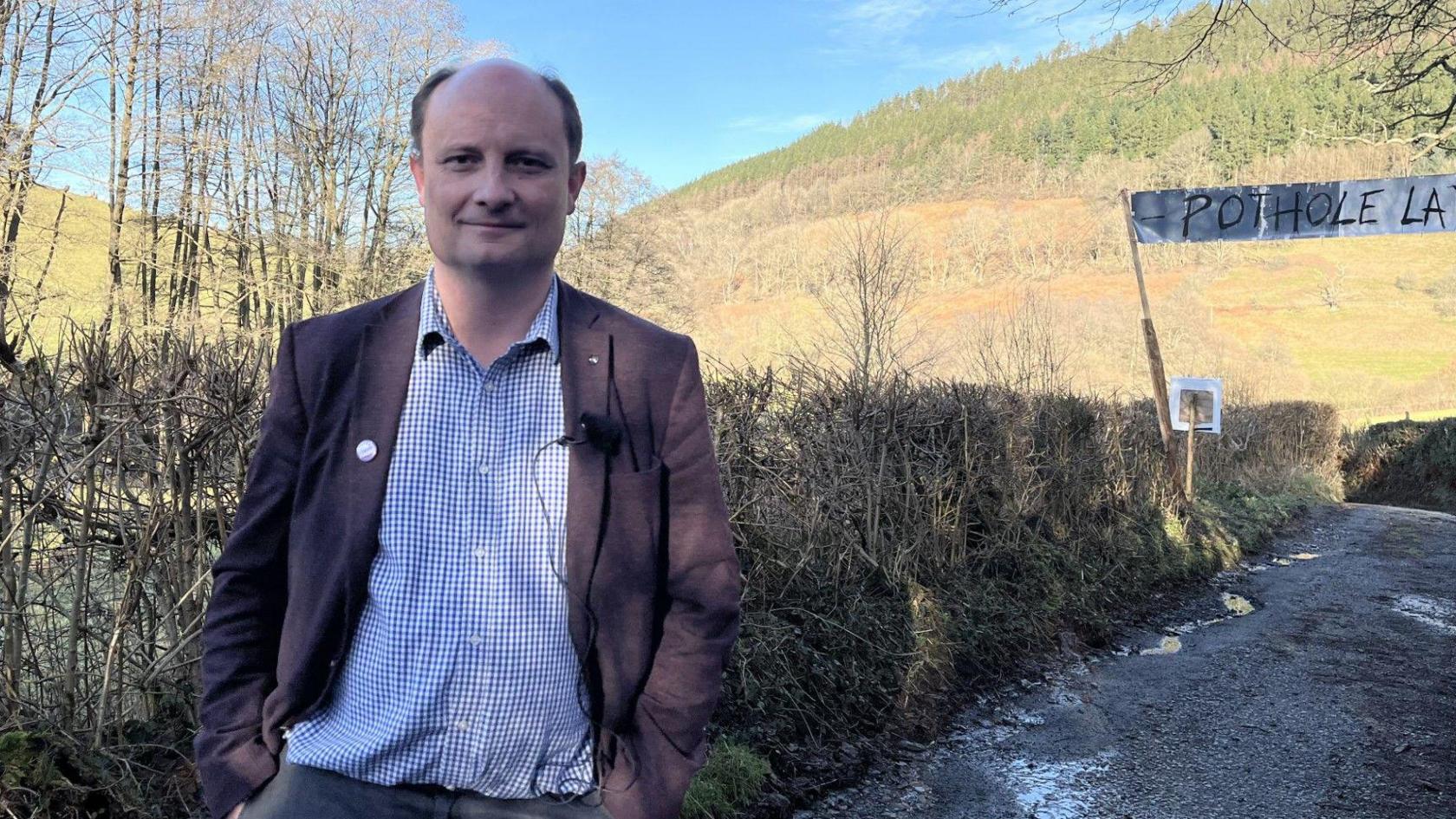 A middle aged man with receding brown hair stands in front of a Pothole Land sign over the country lane. He is wearing a brown blazer, a blue and white check shirt and faded black jeans. 