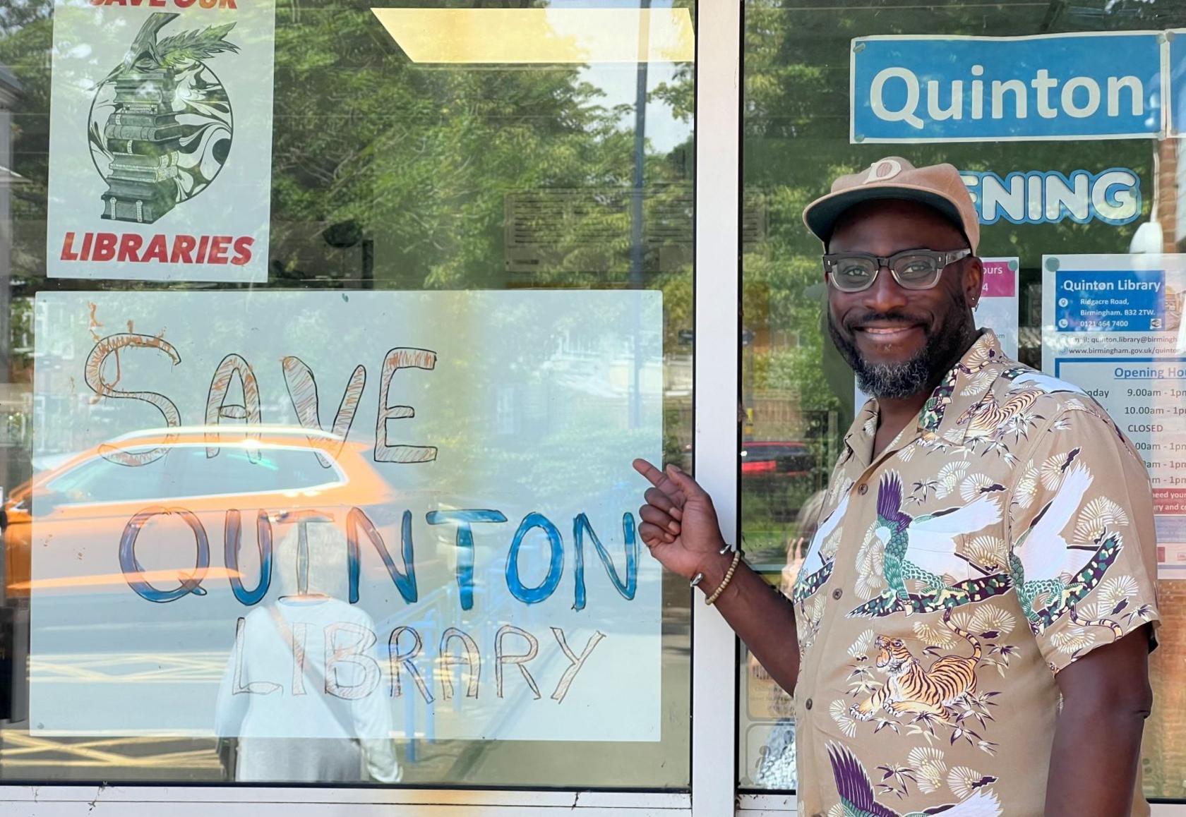 A man wearing a beige shirt and a baseball cap stands by a window and points to a handwritten poster that says "SAVE QUINTON LIBRARY" in capital letters.