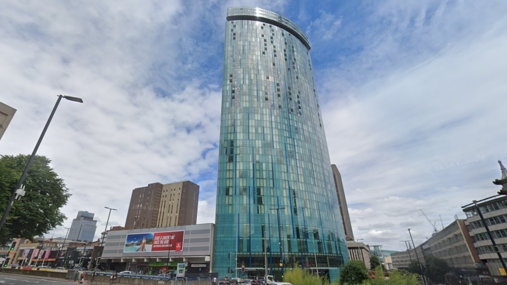 A large skyscraper covered in windows on a busy city street. Other skyscrapers can be seen in the background and cars are on the road below.