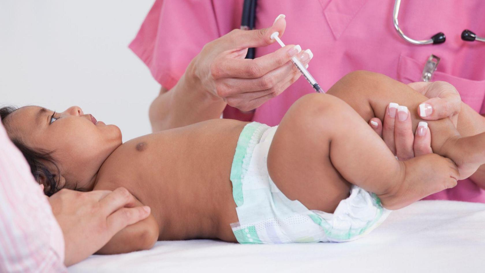 A baby wearing a disposable nappy is lying on her back as a medical practitioner injects her in the leg.