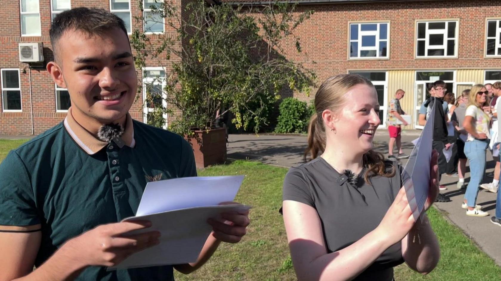 Amen Akhtar wearing a green polo shirt with a fluffy mic clipped to the collar. He is standing beside Hannah Webb, wearing a grey t-shirt and a clip-on mic.  They are both holding sheets of paper with their exam results and smiling.