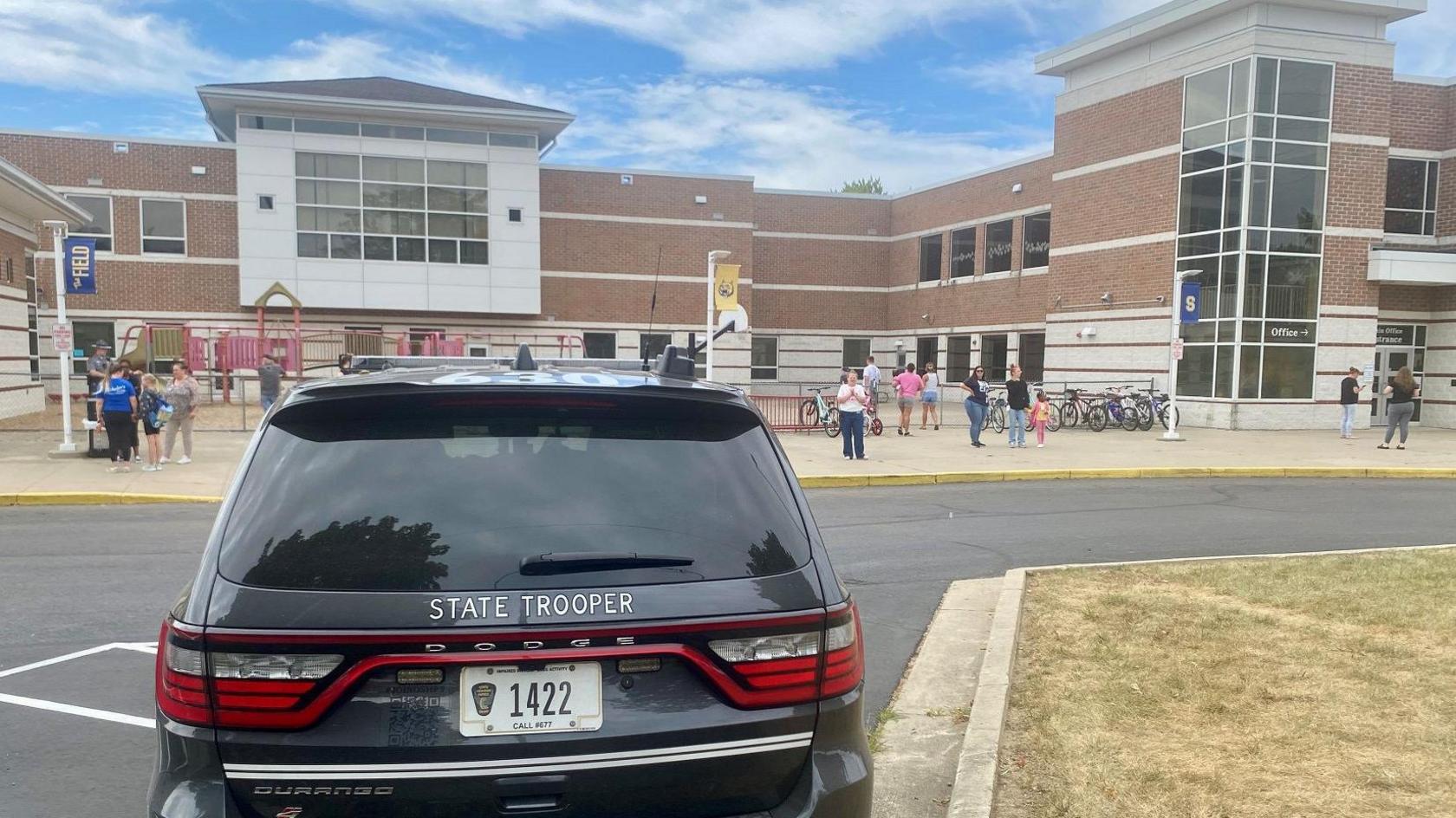 A State Trooper vehicle in the foreground, with a school and children outside of it in the background