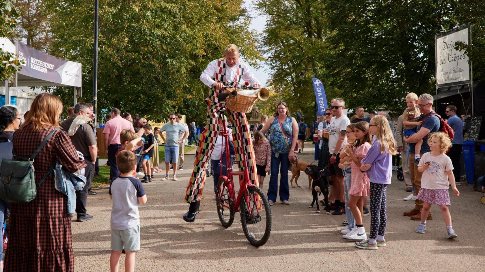 A circus performer wearing long patchwork trousers, on stilts, on an abnormally large red bicycle that has a horn on its basket, surrounded by families and trees, on a park pathway