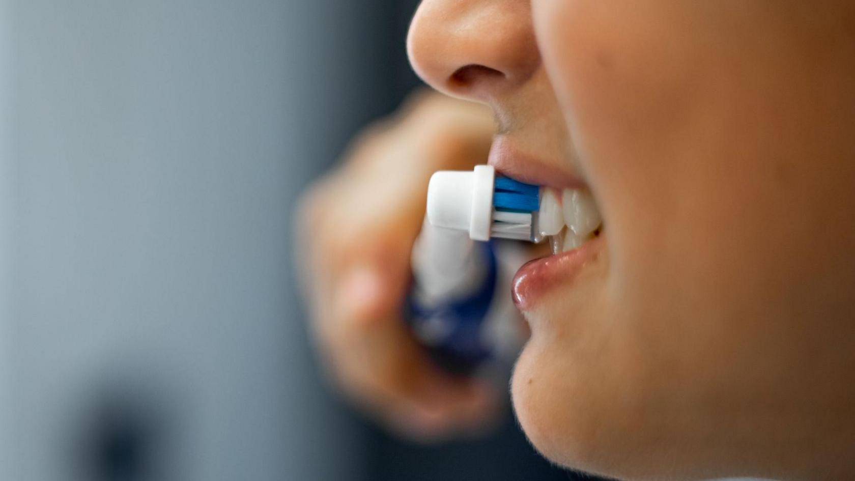 A close-up stock picture of the mouth of a teenage boy who is holding an electric toothbrush and brushing his teeth.