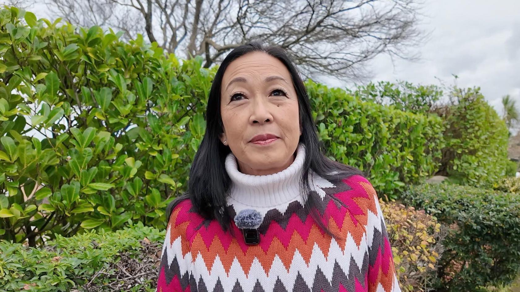 A woman in a colourful patterned jumper stands in a residential street with hedges in the background. 