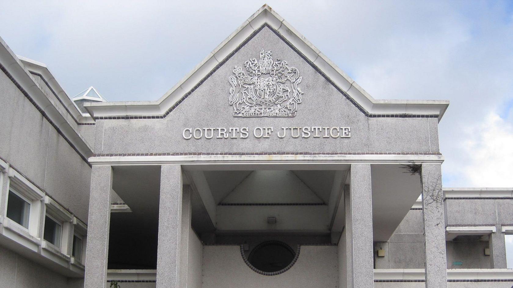 The front of Truro Crown Court. It is grey stone building with COURTS OF JUSTICE written below an elaborate heraldic shield. There are four pillars and the roof is pitched to a point.