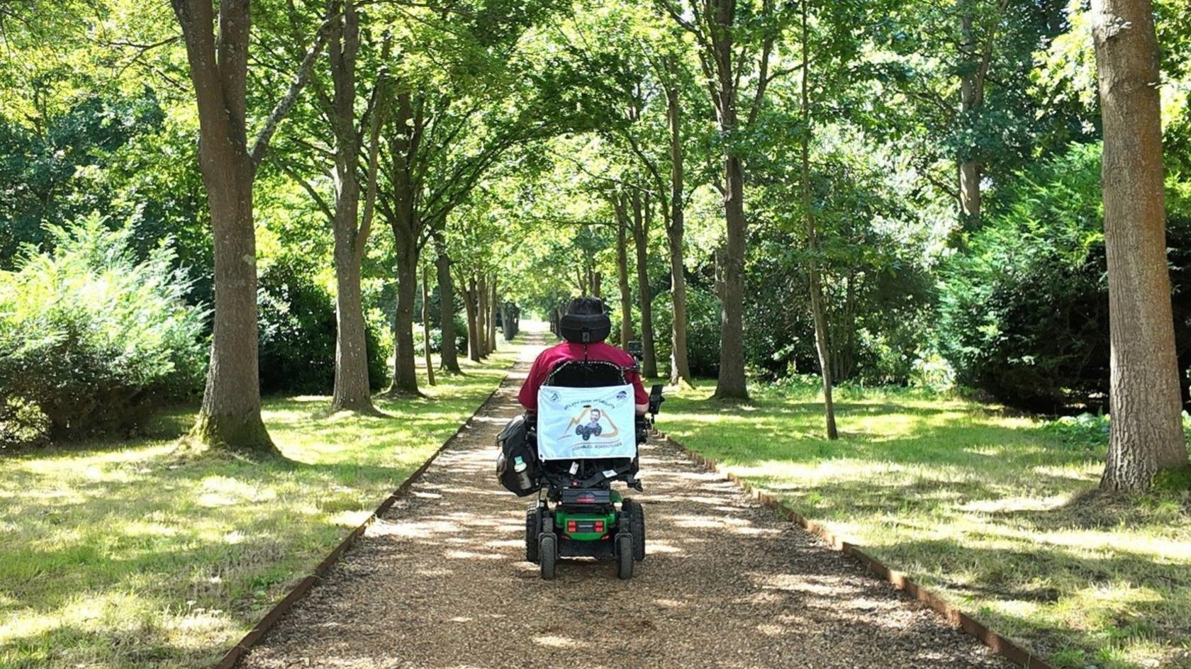 A man in a wheelchair moves away from the camera along a gravel path with grass and trees on either side.