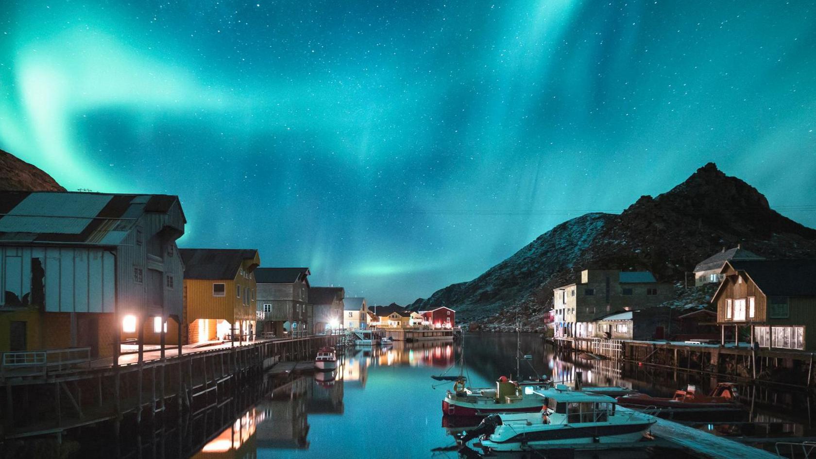 Light blue lights inthe sky above a village. Small boats are on a river and buildings line the water. There is a rocky mountain in the background.